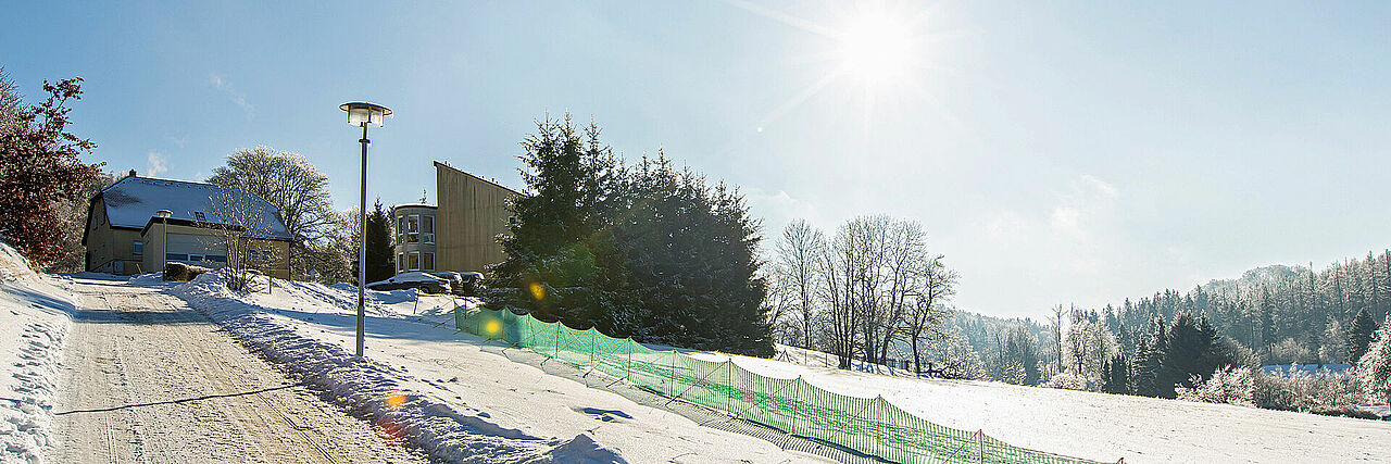 Waldweg mit Blick zum Gebäude der Bergwacht bei Schnee, Aufnahme im Gegenlicht