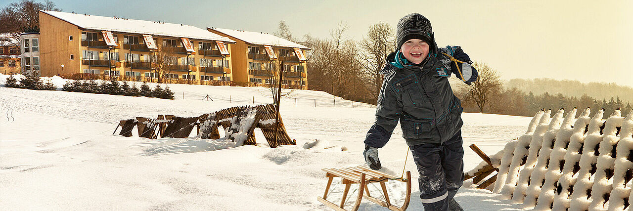 Kind mit Schlitten, im Hintergrund Bettenhaus der Kurklinik Haus am Jonsberg im Winter