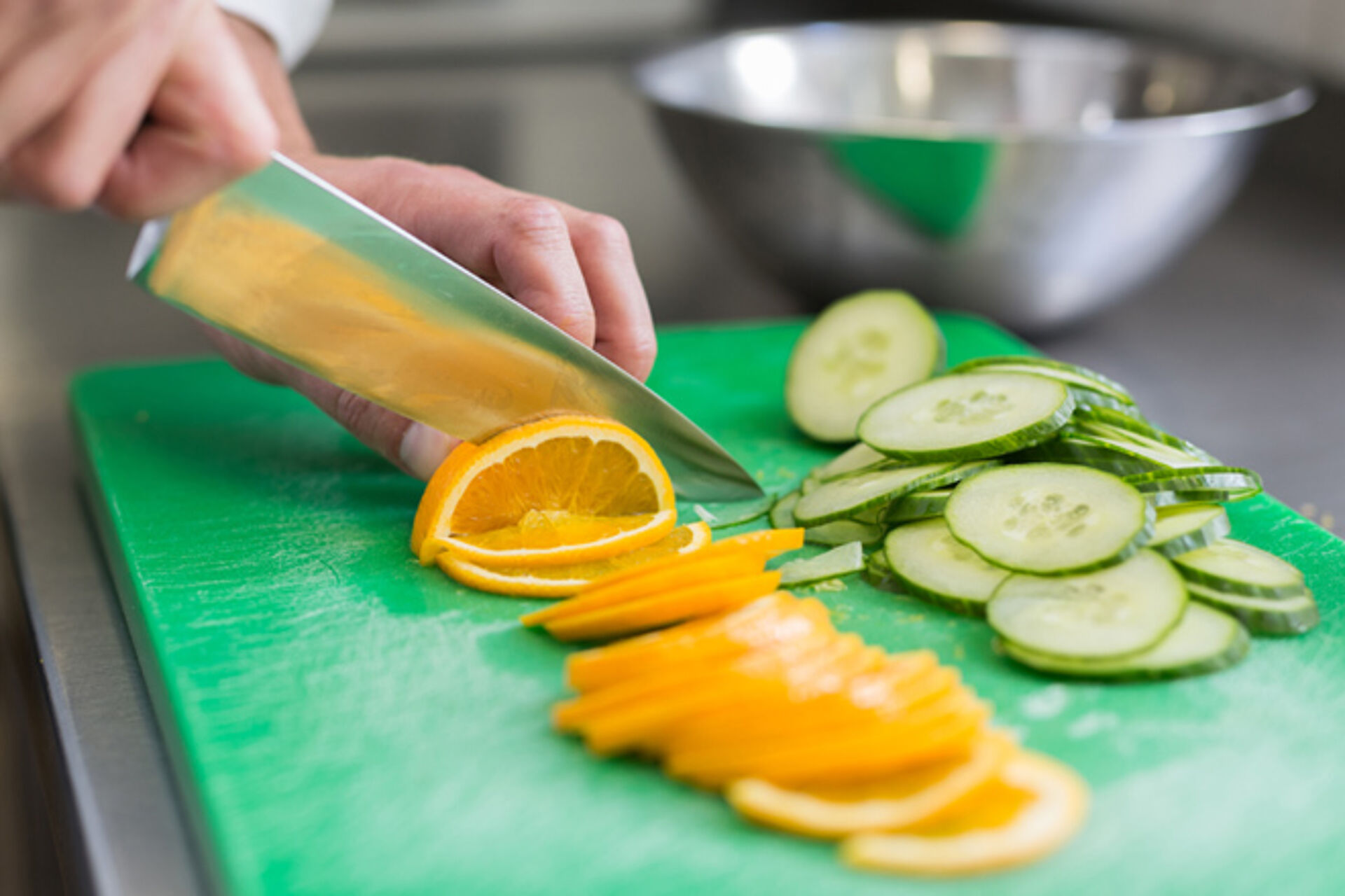Zubereitung Detoxwasser: Grünes Schneidbrett mit Orangen und Gurkenscheiben, Hand führt großes Schneidmesser