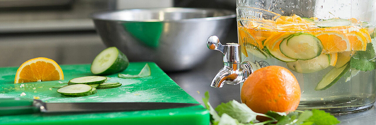 1920_Detoxwasser1.jpg Küchenutensilien Schüssel, Messer und Schneidbrett sowie Wasserkaraffe mit Gurken und Orangen