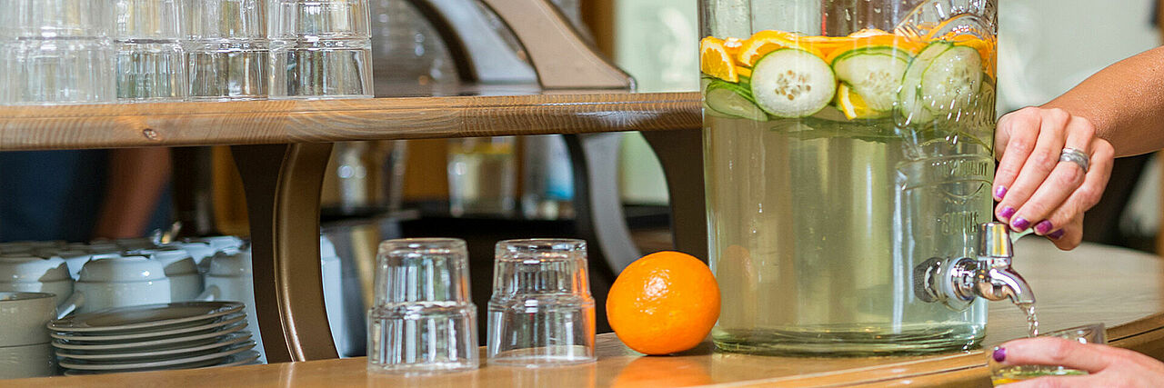 1920_Detoxwasser.jpg Karaffe mit Gurken-Orangen-Detoxwasser, Wassergläser und Hand, die gerade Wasser aus der Karaffe lässt