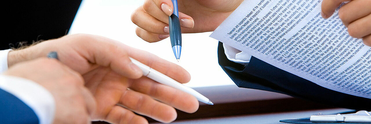 Dekofoto mit den Händen von zwei Personen, beide halten einen Stift, eine Person hält zusätzlich ein Blatt Papier