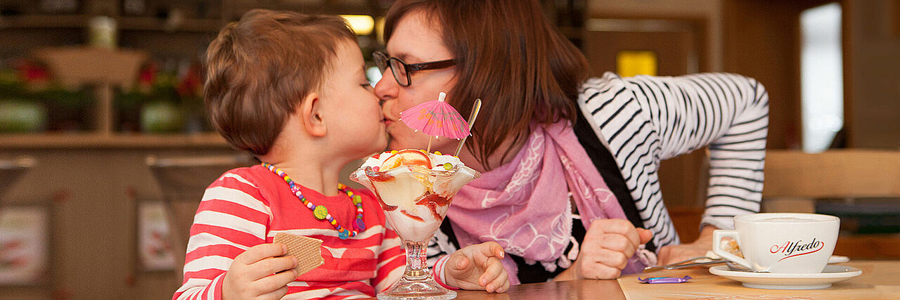 Mutti küsst Kind auf den Mund, im Vordergrund stehen ein Eisbecher und eine Tasse Kaffee